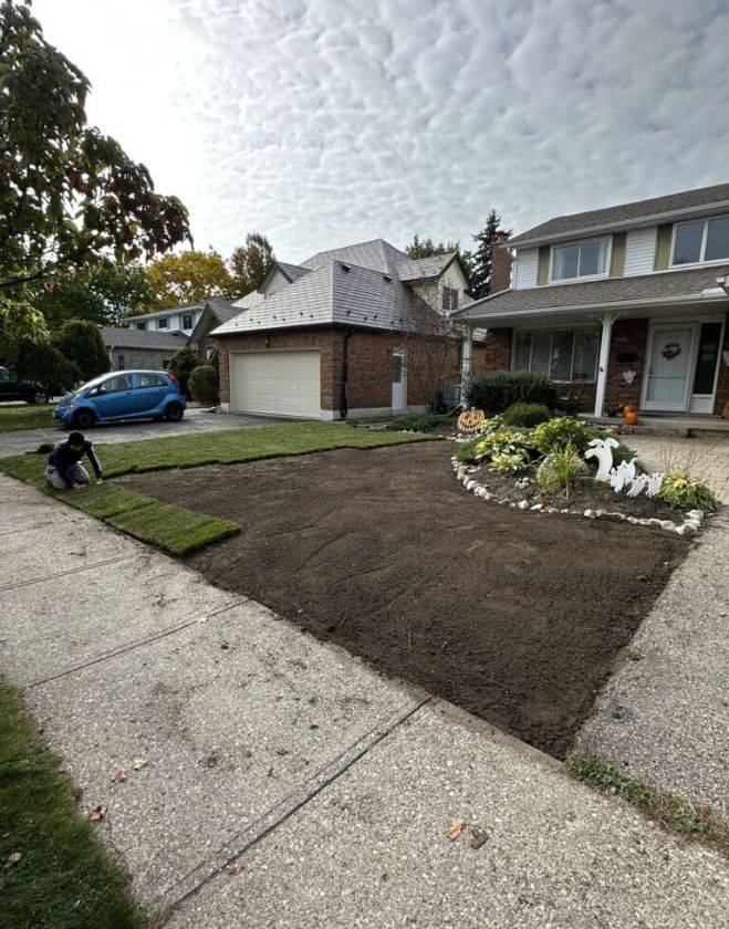 A professional landscaper kneeling to lay fresh green sod rolls on precisely graded dark topsoil in a residential front yard in Hamilton, Ontario.