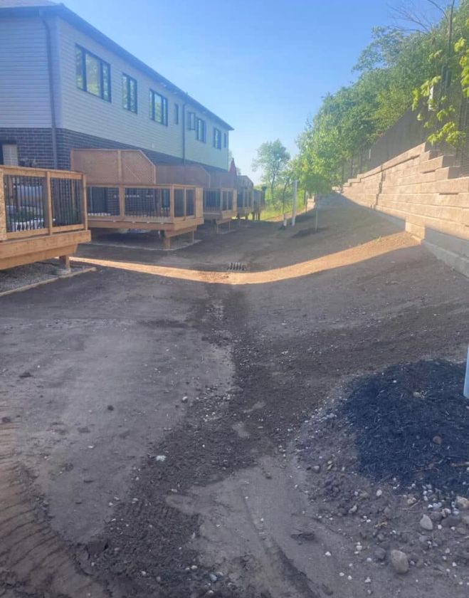 Graded bare soil shaped perfectly around a drainage swale behind wooden decks in a Hamilton residential complex, prepared for grub treatment and fresh sod.