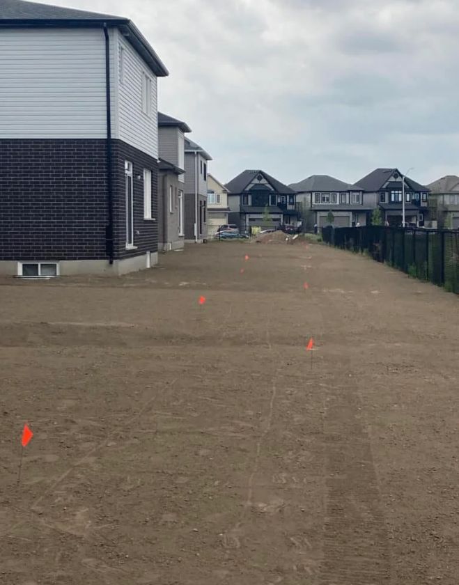A large expanse of freshly leveled bare dirt beside a newly built brick home in a Hamilton subdivision, marked with orange utility flags after professional rough and final grading.