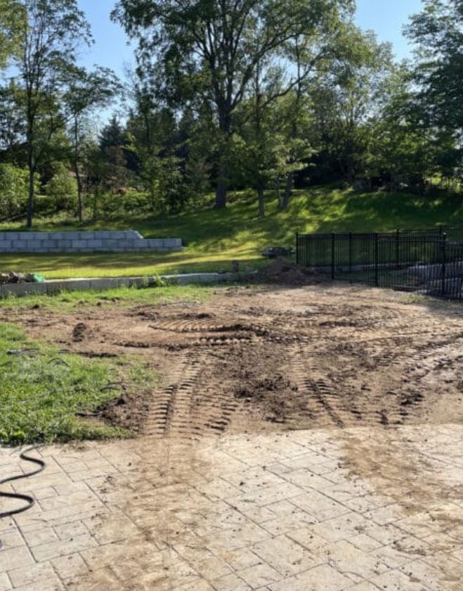 Lawn Repair 4 A residential backyard in Hamilton with damaged grass removed, showing bare dirt with equipment tracks next to a stone patio and a black metal fence.